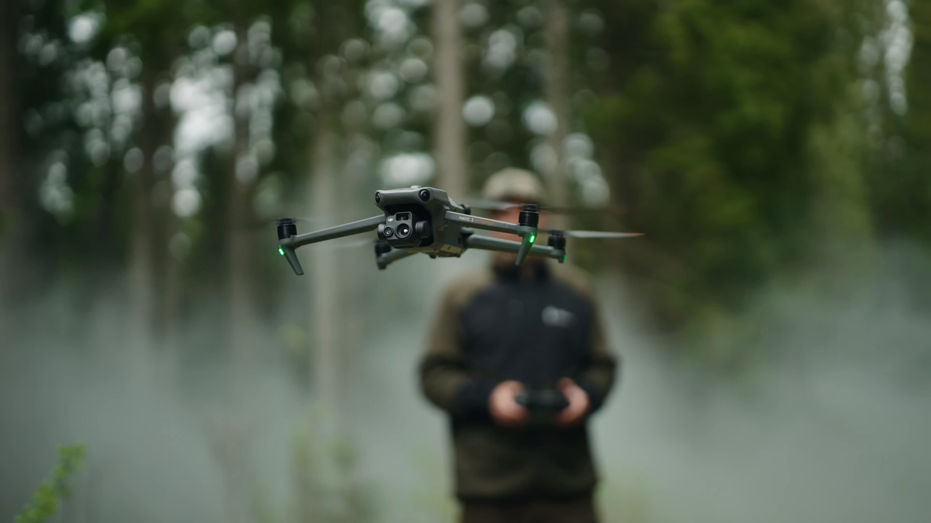 En drönare svävar i luften framför en person som håller en fjärrkontroll i en dimmig skogsmiljö.