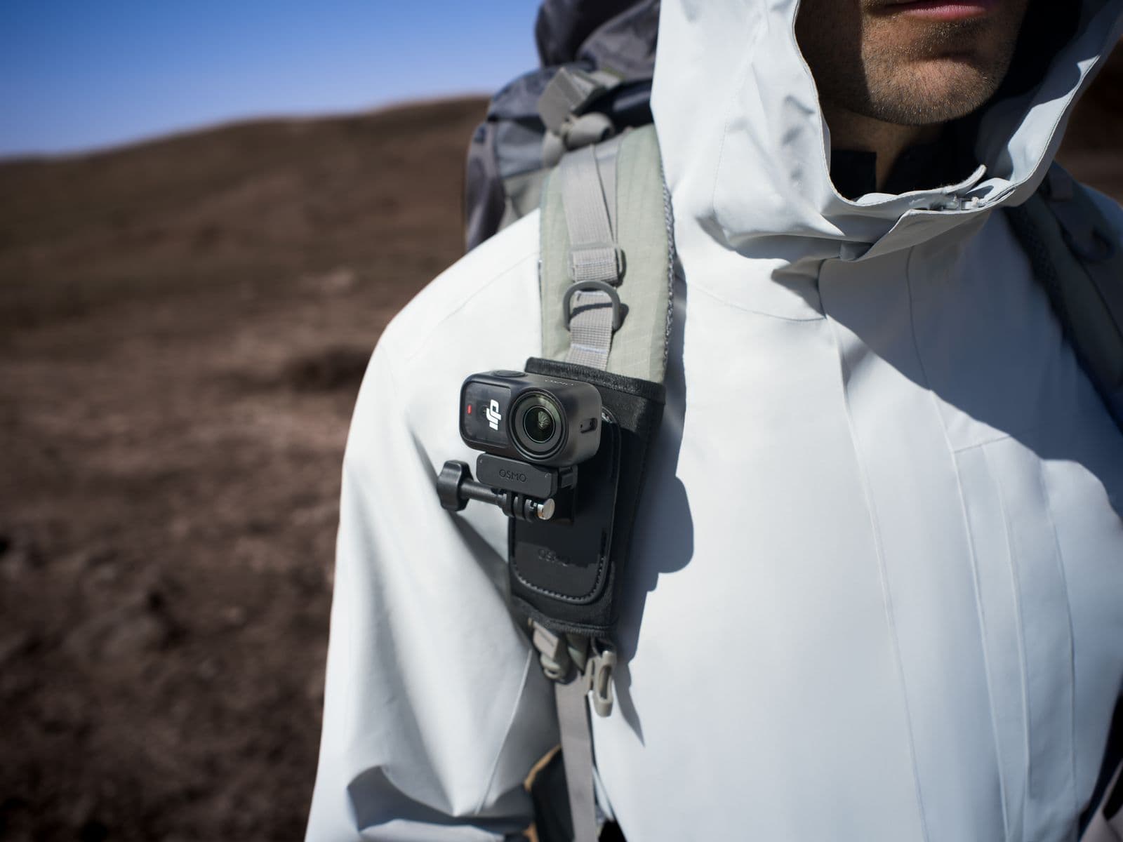 En person i en vit jacka bär en DJI Osmo Nano fäst på ryggsäckens axelrem, redo för äventyr i ett kargt landskap.