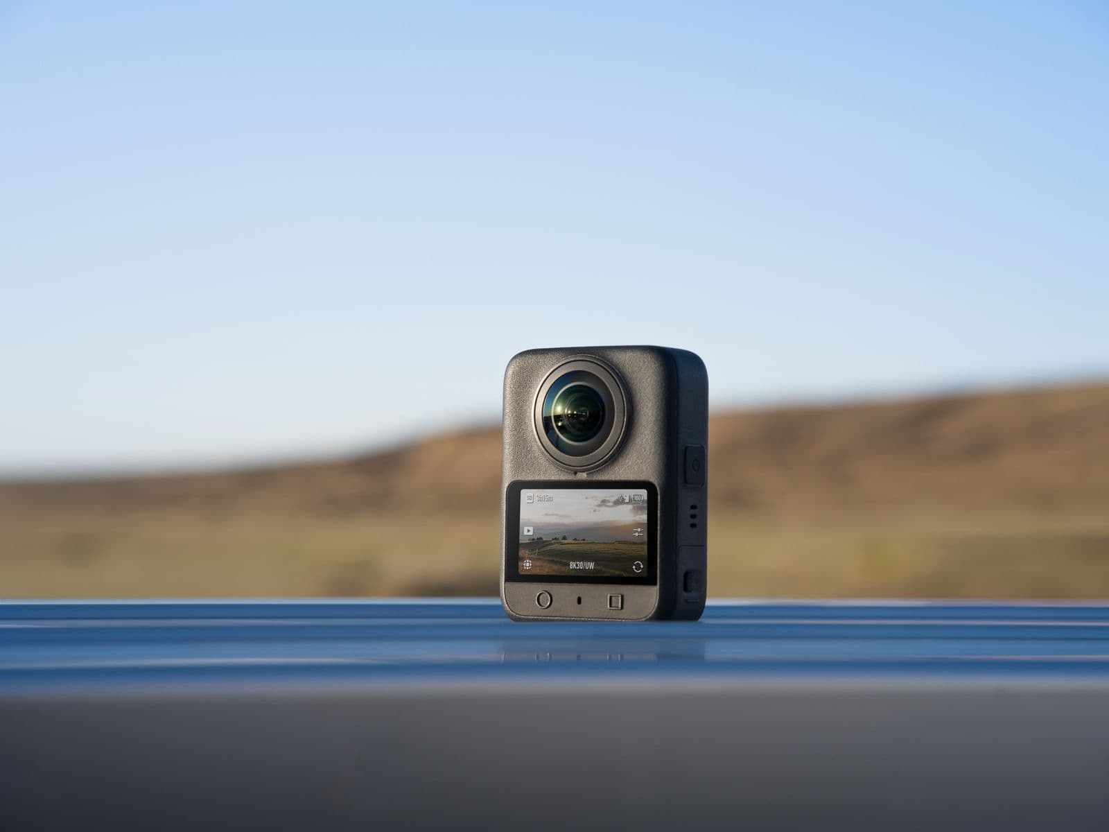 En actionkamera står på en reflekterande yta med en suddig landskapsbakgrund under en klar himmel.