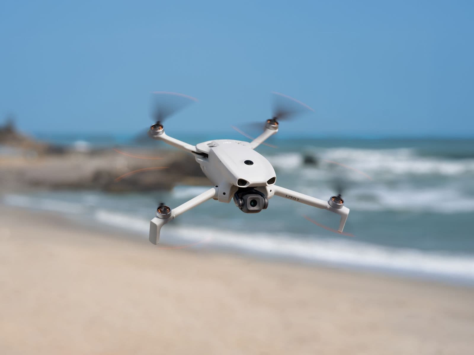 En vit drönare svävar över en sandstrand med havet i bakgrunden.