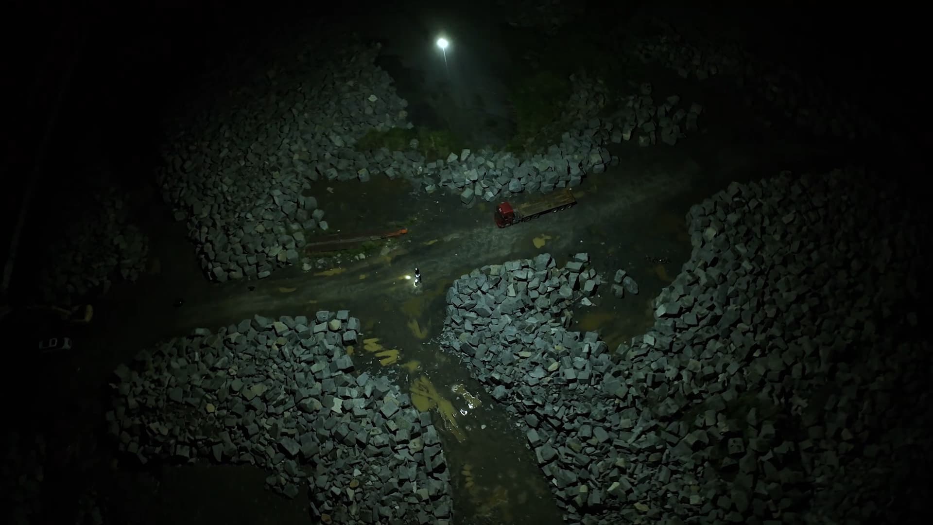 En nattlig scen med en person som går längs en grusväg, omgiven av stora stenblock, upplyst av en CZ100 i luften.