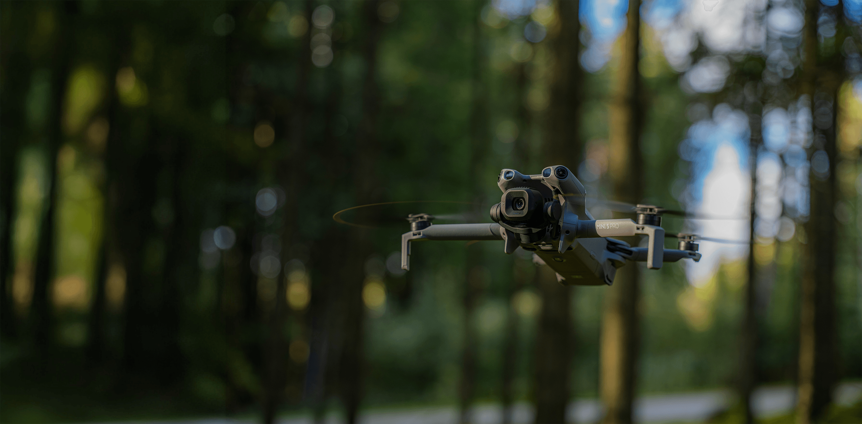 En DJI Mini 5 Pro-drönare svävar genom en skogsmiljö, omgiven av höga träd och mjukt ljus.