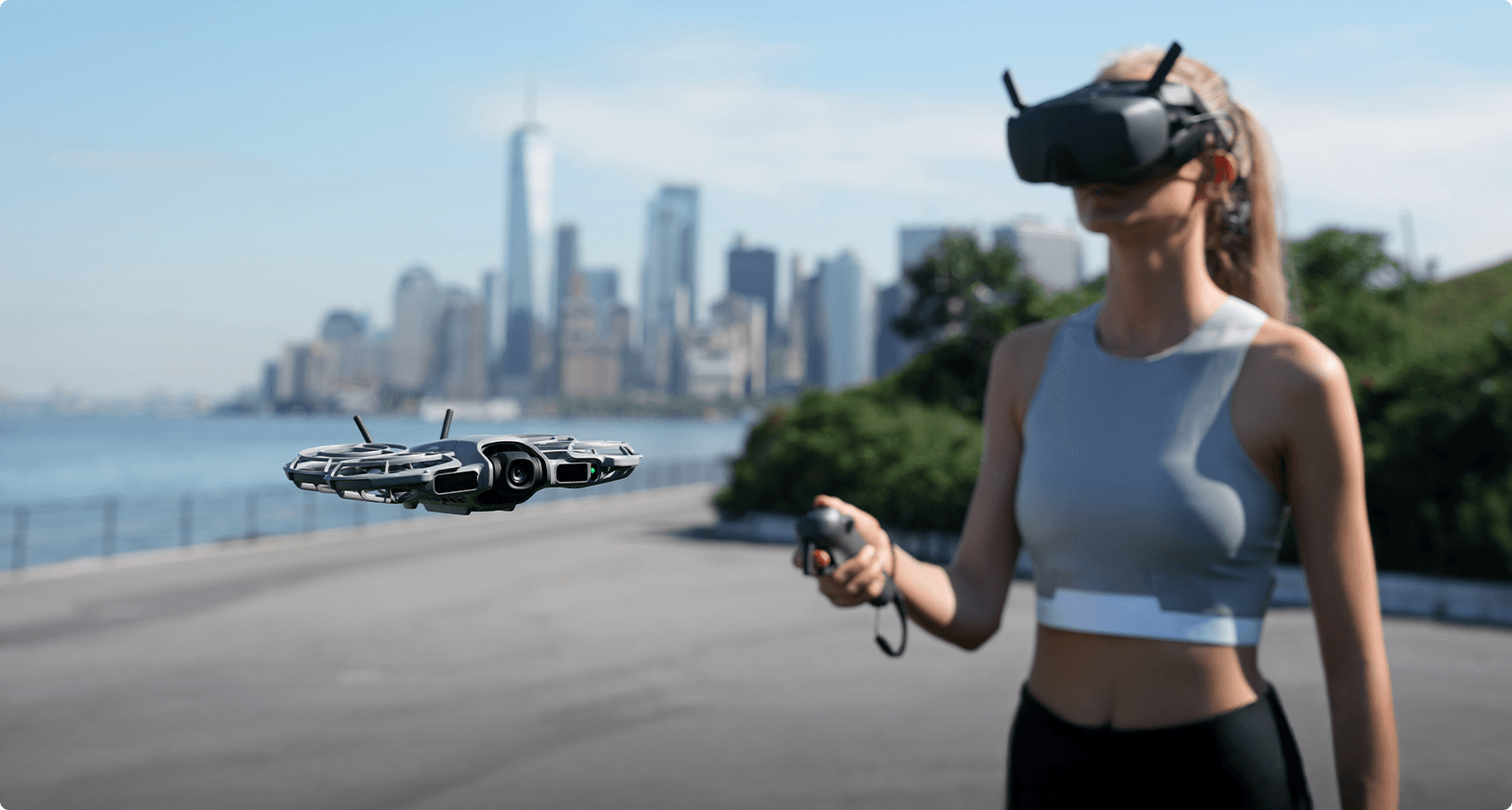 En person med VR-headset styr en DJI Neo 2-drönare vid en vattensida med en stadssilhuett i bakgrunden.