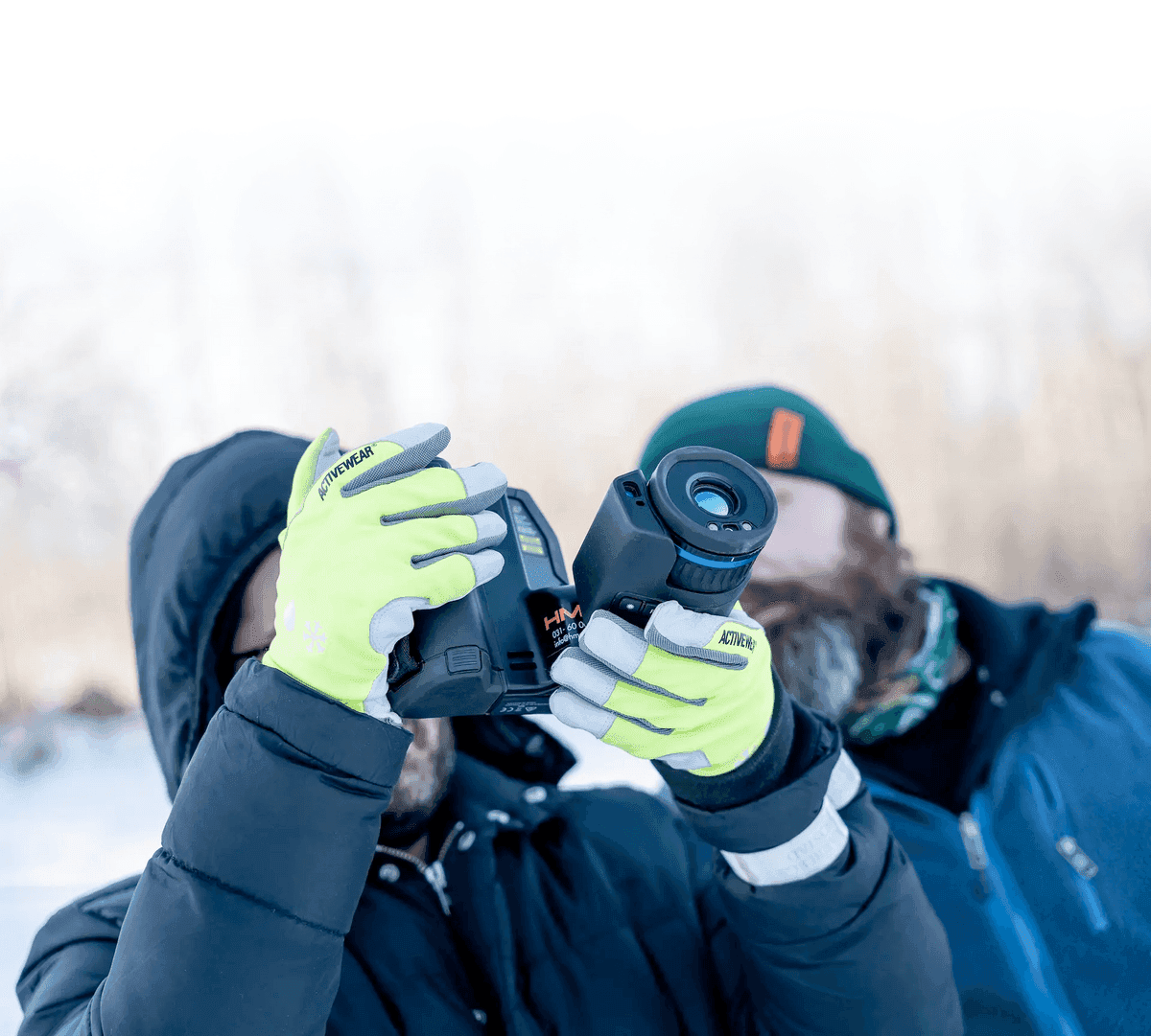 Två personer i vinterkläder använder en handhållen termisk kamera utomhus.