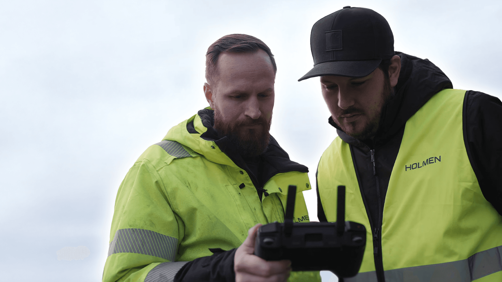 Två personer i gula säkerhetsvästar styr en drönare med en fjärrkontroll, medan drönaren svävar i luften ovanför.