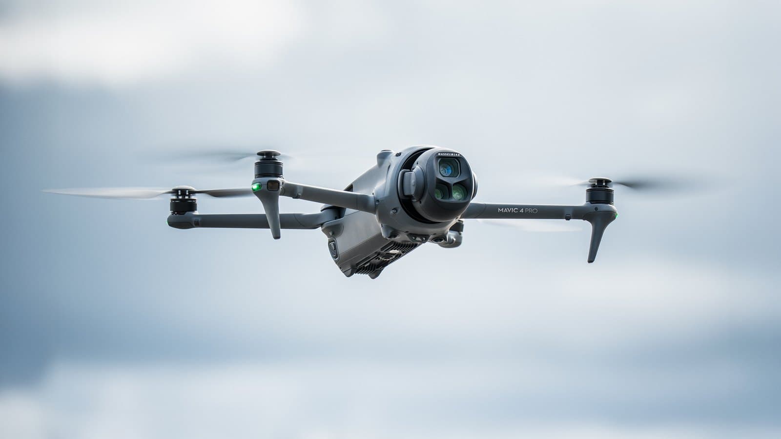 En DJI Mavic 4 Pro-drönare svävar i luften mot en molnig himmel, med sina rotorblad i rörelse.
