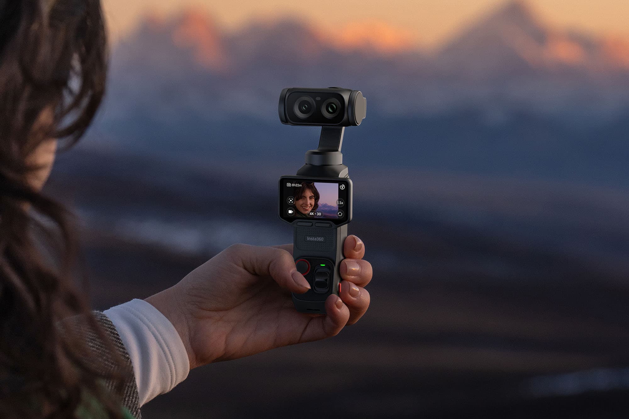 En person håller en kompakt kamera med en skärm som visar en selfie, mot en bakgrund av suddiga berg i solnedgången.