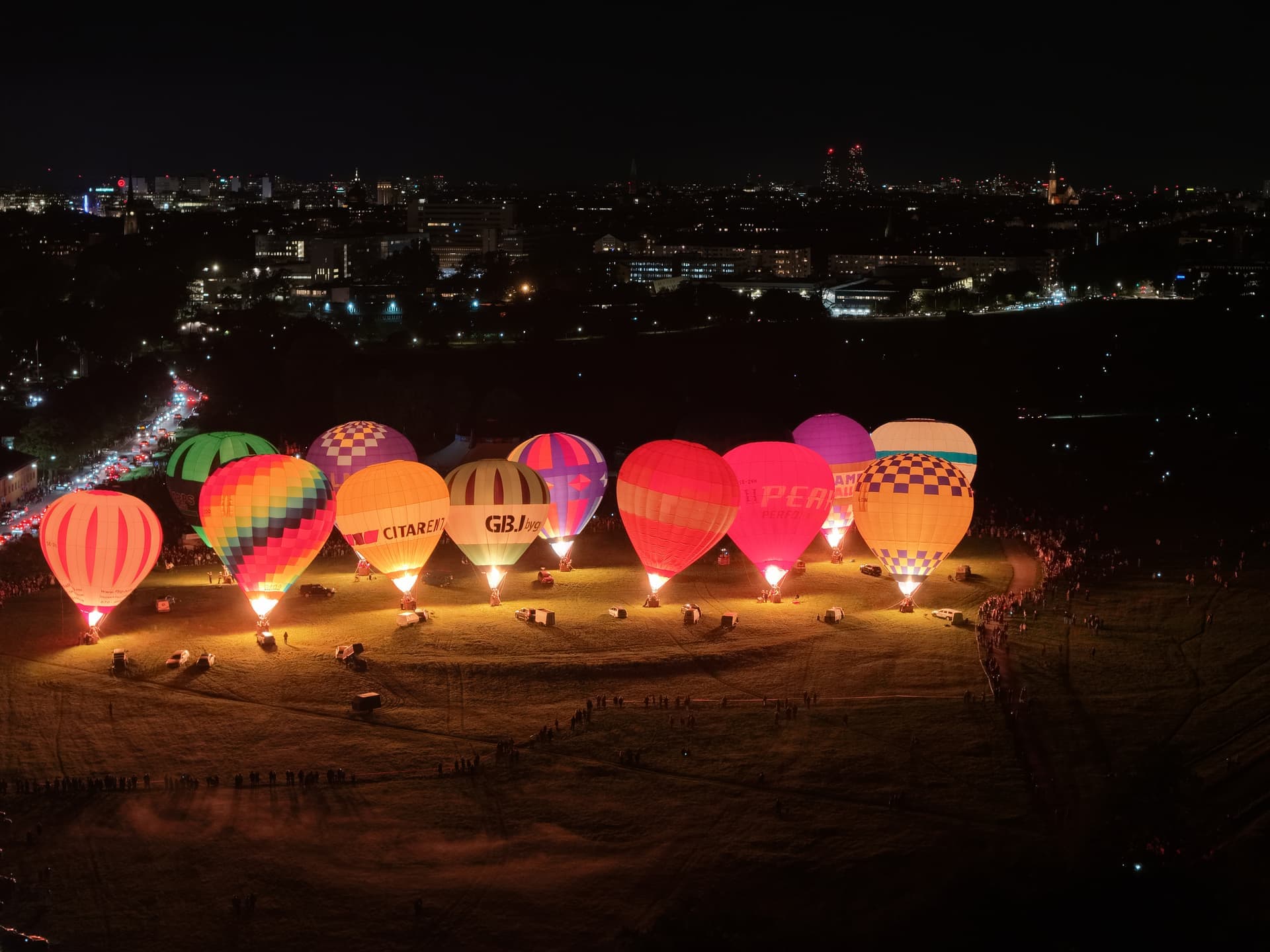 Flera färgglada luftballonger lyser upp en nattlig himmel över en stad, omgivna av åskådare på marken.