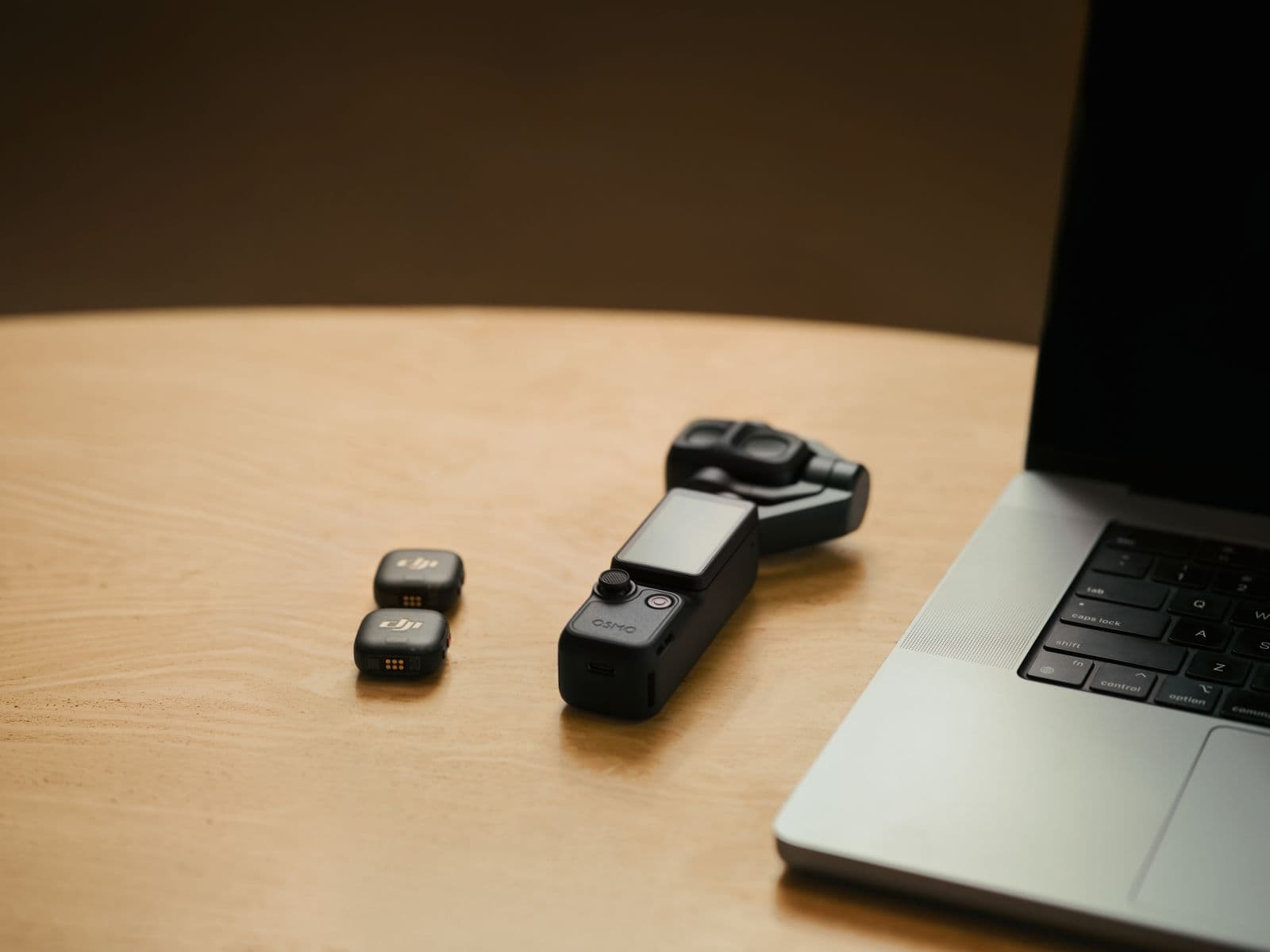 En kompakt kamera och två små enheter ligger bredvid en bärbar dator på ett träbord, vilket skapar en modern och teknisk arbetsyta.