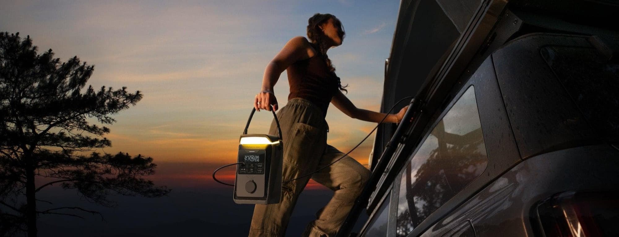 En person klättrar upp på en bil med en bärbar kraftstation i handen, mot en bakgrund av en färgglad solnedgång.