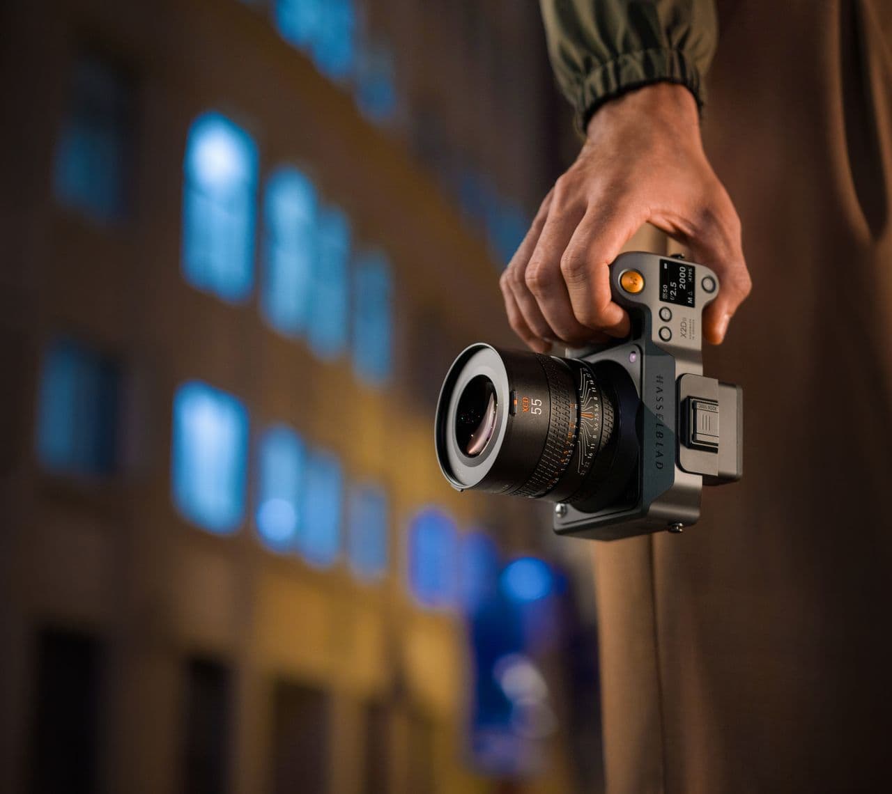 En person håller en Hasselblad X2D II 100C kamera i handen, med en suddig stadsmiljö i bakgrunden.