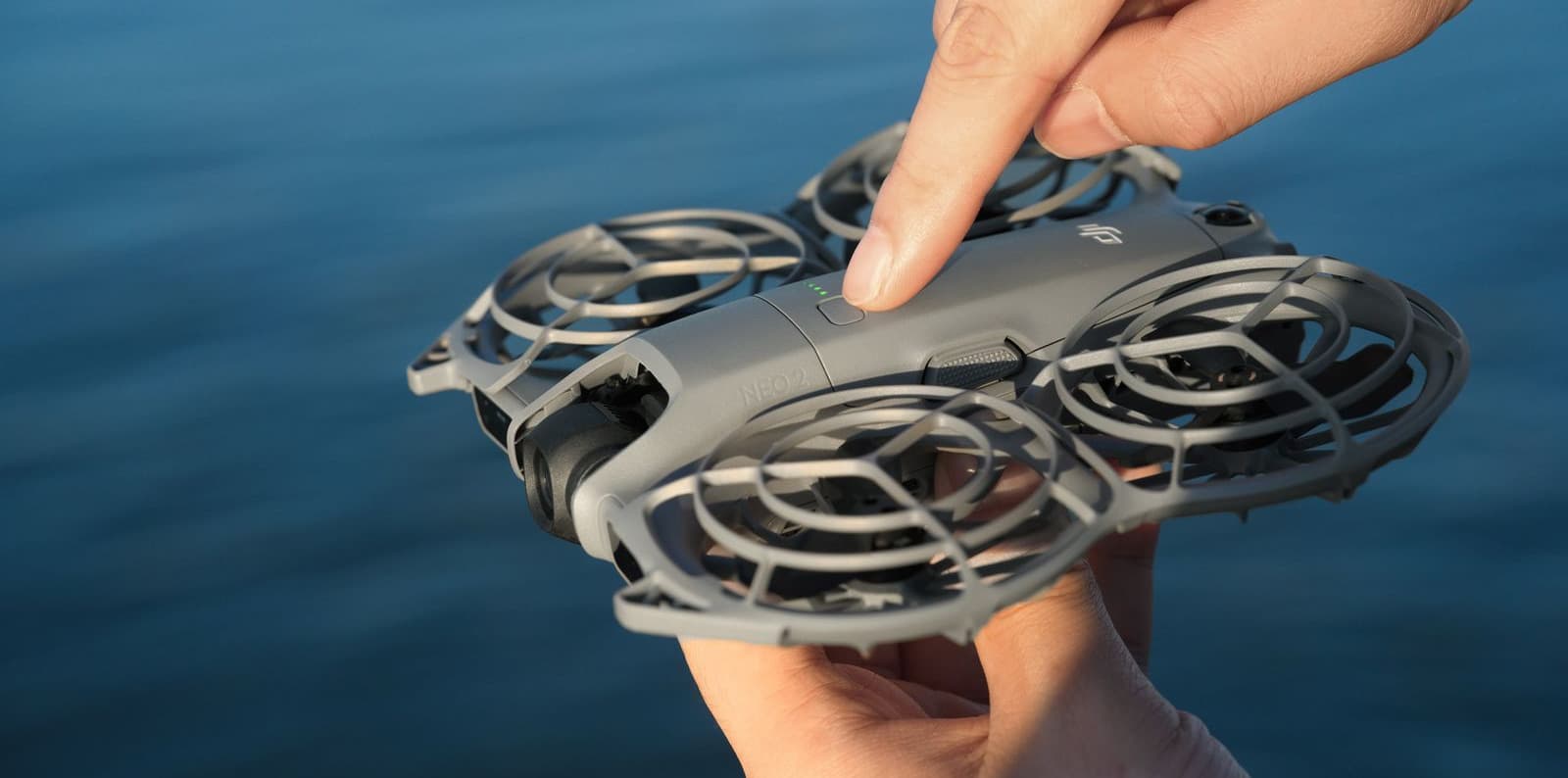 En person håller en kompakt DJI Neo 2-drönare över vatten och trycker på en knapp för att aktivera den.