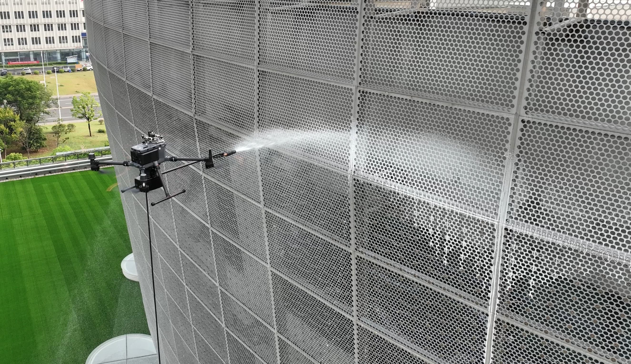 En drönare rengör en perforerad fasad på en byggnad med en vattenstråle, ovanför en grönskande gräsmatta.