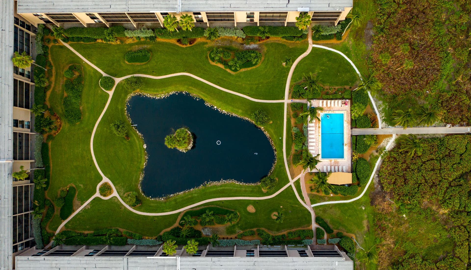 Flygbild av en symmetrisk trädgård med en central damm och en pool omgiven av grönska och gångvägar.