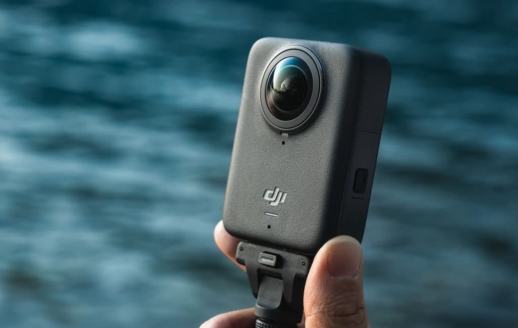 En hand håller en DJI Osmo 360 kamera vid en sjö, med vattnet suddigt i bakgrunden.