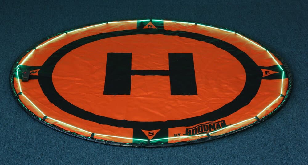 En orange Hoodman landningsplatta för drönar med ett stort 'H' i mitten, omgiven av lysande gröna ljus, placerad på en mörk yta.