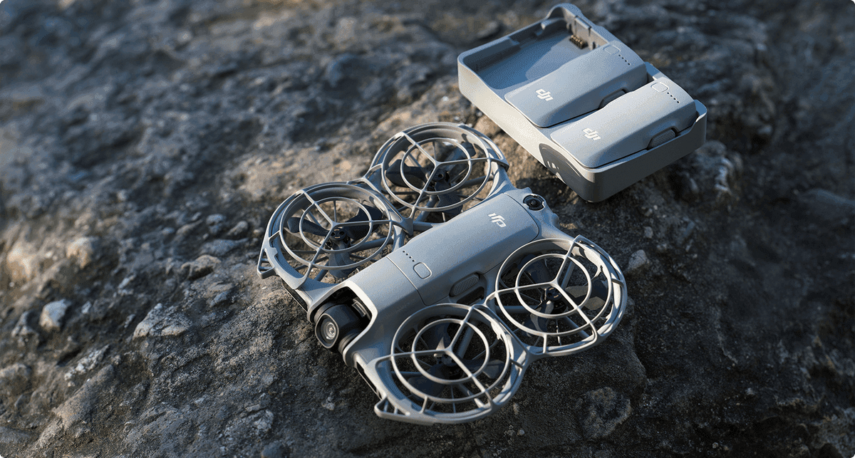 En kompakt DJI Neo 2-drönare med skyddade propellrar ligger på en stenig yta bredvid ett laddningsetui.