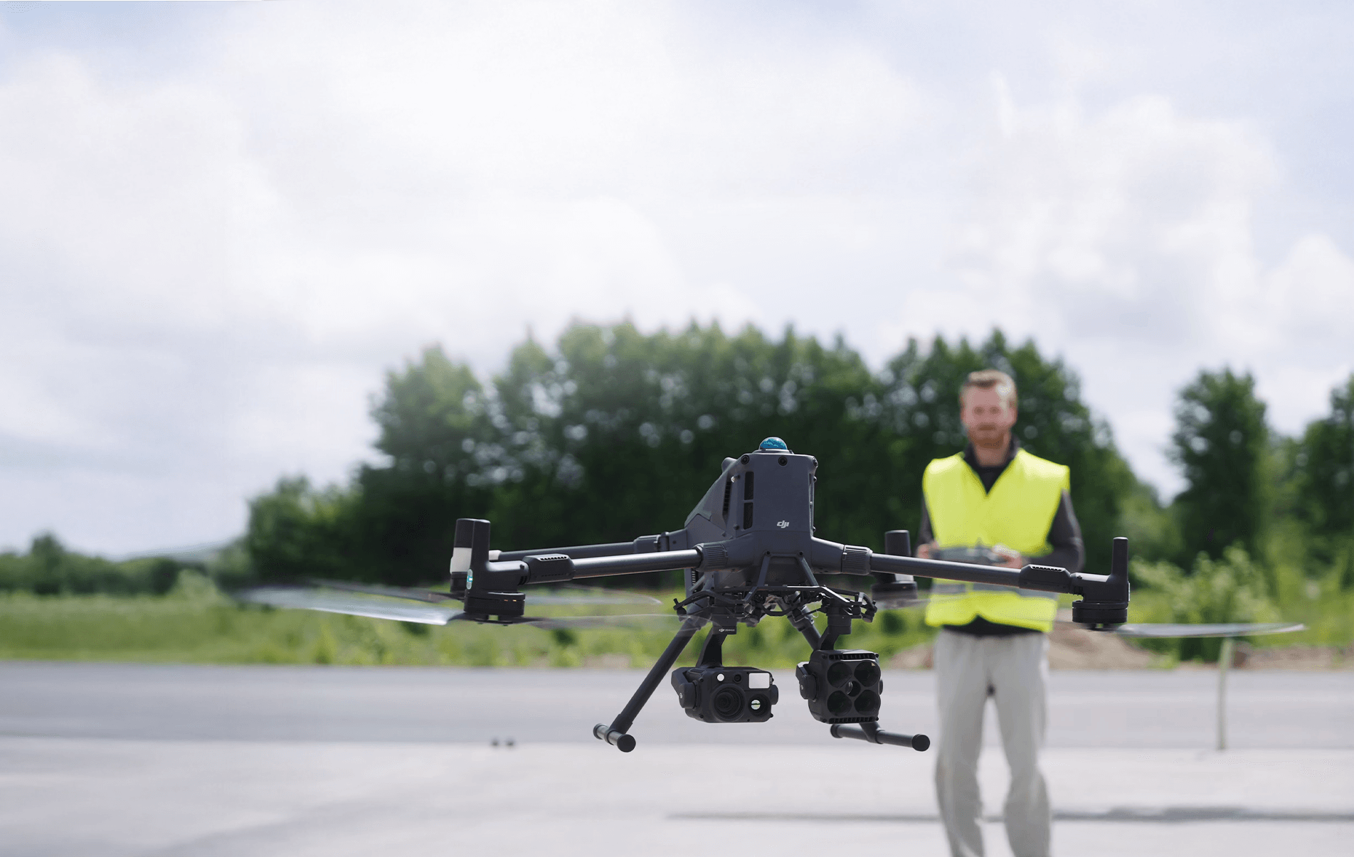 En Matrice 400 svävar i luften medan en drönarpilot i gul väst styr den med en fjärrkontroll i en utomhusmiljö.