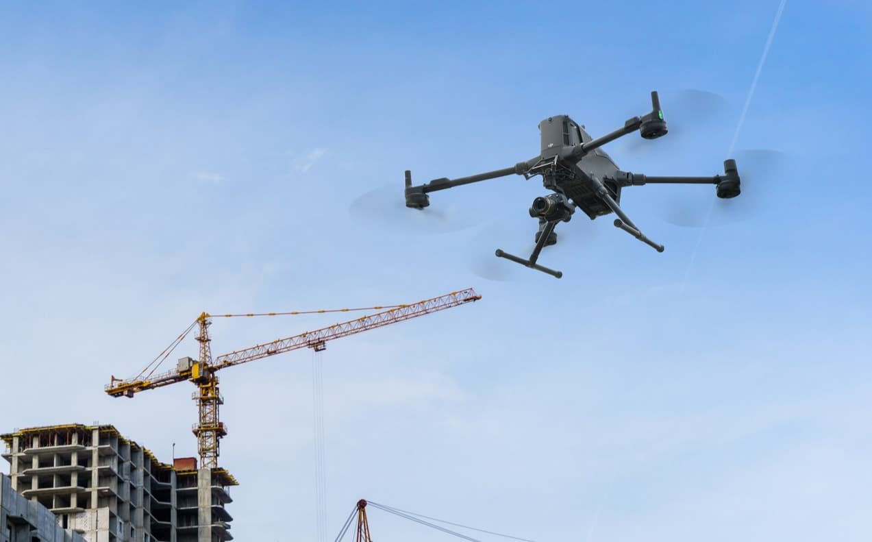 En drönare svävar över en byggarbetsplats med höga kranar och halvfärdiga byggnader mot en klarblå himmel.