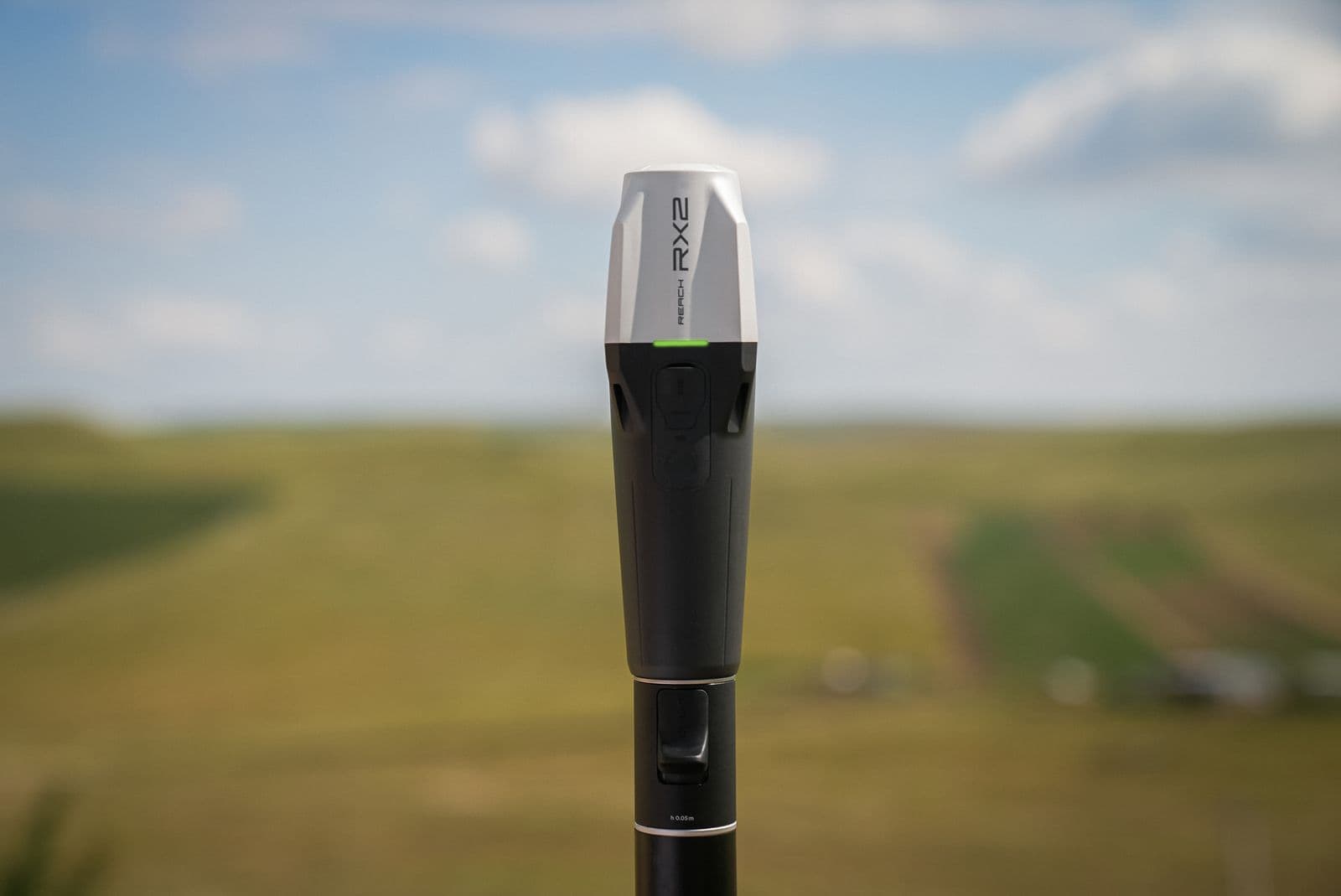 En modern Emlid Reach RX2 står upprätt mot en suddig landskapsbakgrund under en klar himmel.