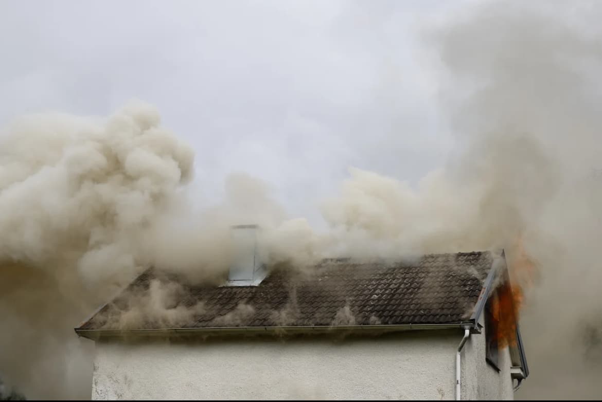 Rök väller ut från taket på ett hus i brand, med lågor synliga vid sidan.