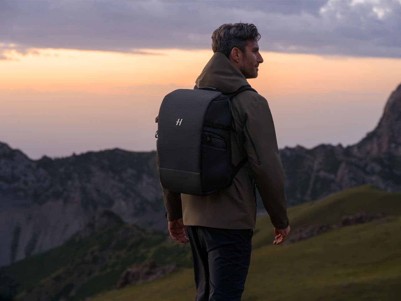 En person med en ryggsäck står på en bergstopp och blickar ut över en naturskön vy vid solnedgången.
