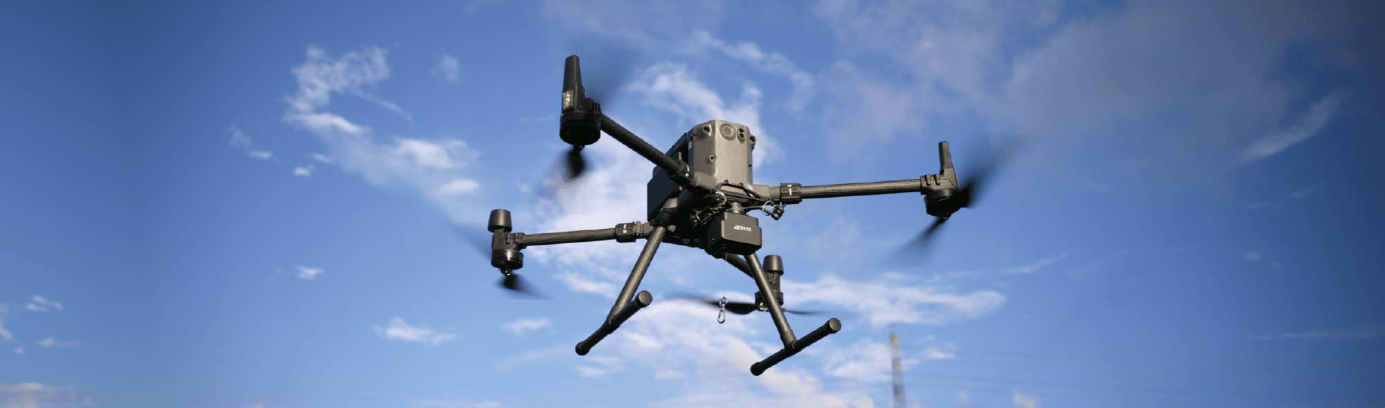 En drönare svävar i luften mot en klarblå himmel, med propellrarna i rörelse.