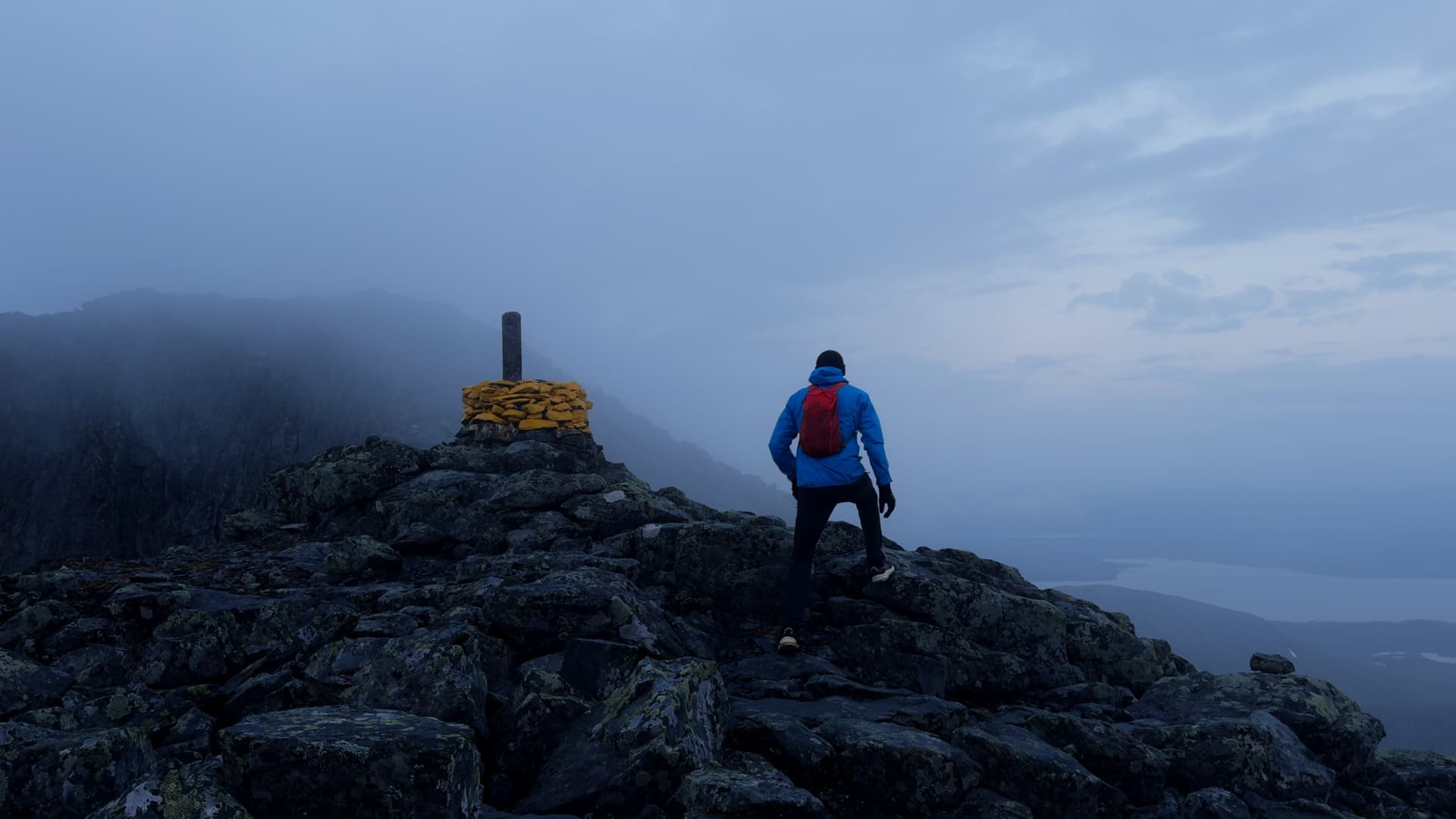 En person i blå jacka och röd ryggsäck vandrar uppför en stenig bergstopp i dimmigt väder.