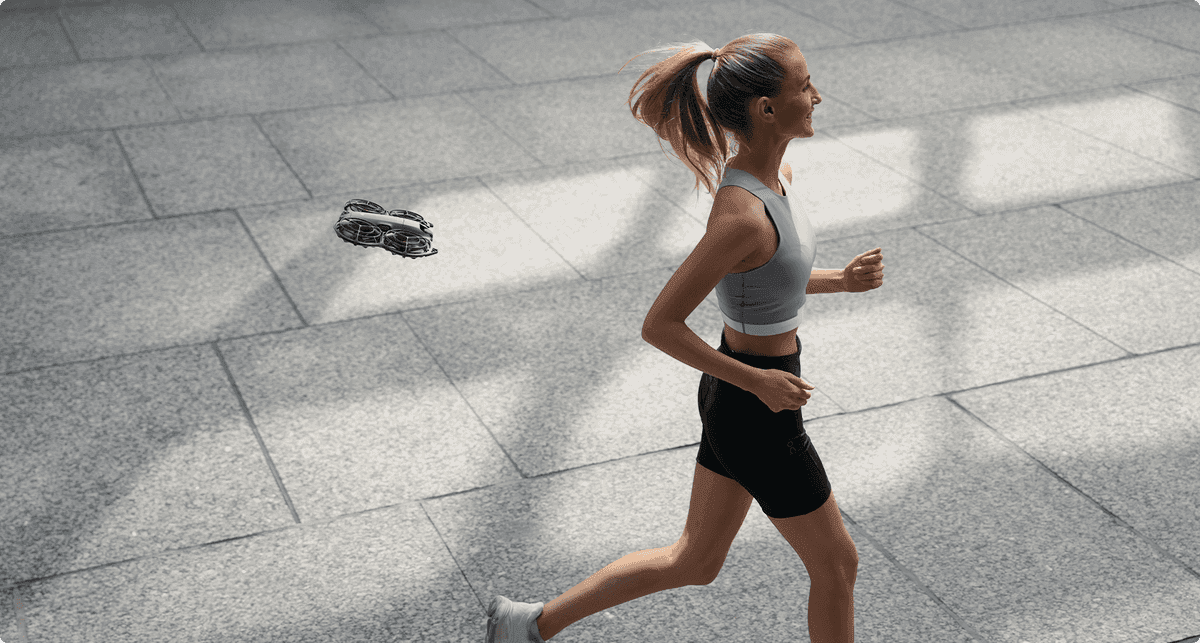 En kvinna joggar på en solbelyst trottoar medan en liten DJI Neo 2-drönare svävar bredvid henne.