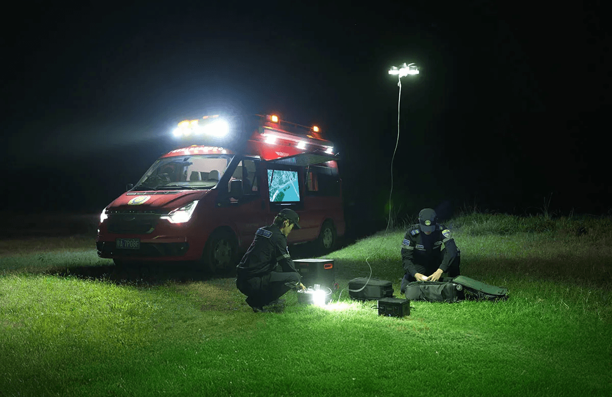 Två personer arbetar med utrustning under en CZ10 Tethered Hover Light som lyser upp gräsytan bredvid ett rött utryckningsfordon under natten.