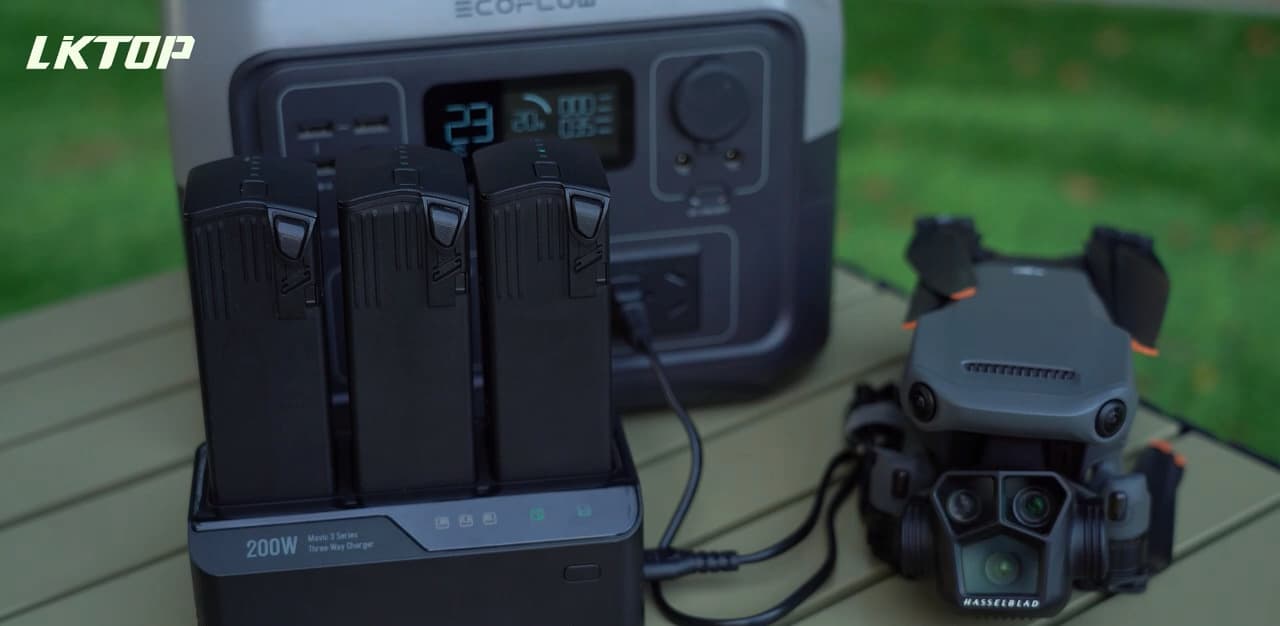 A charging station with three batteries is connected to a power unit, alongside a compact drone on a table.