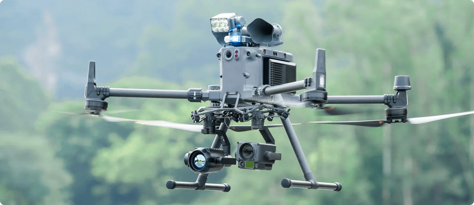 En avancerad DJI Matrice 350-drönare med flera kameror och sensorer från CZI svävar i luften mot en bakgrund av grönskande träd.