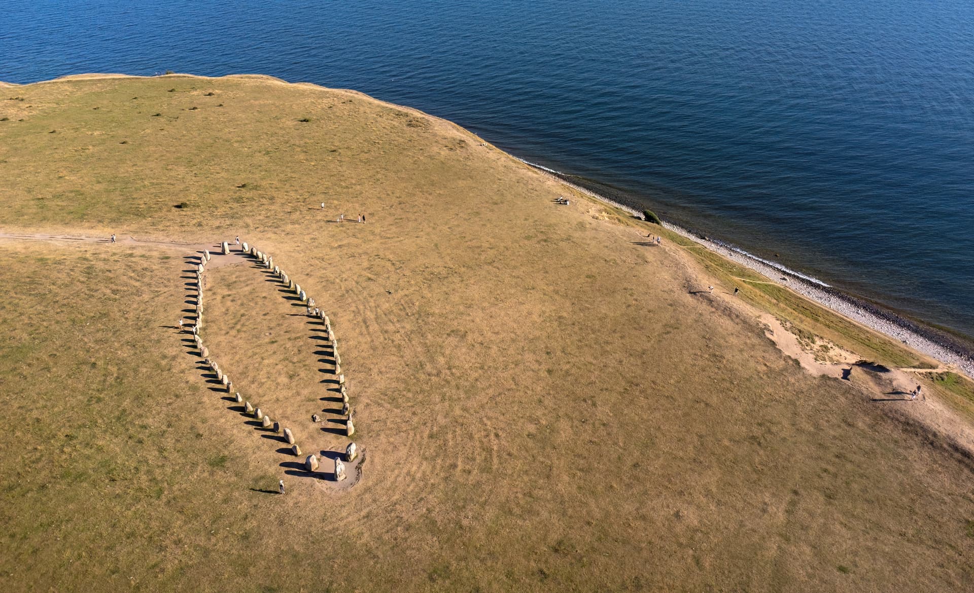 En stensättning formad som ett skepp ligger på en gräsklädd udde vid havet, med några människor som promenerar i närheten.