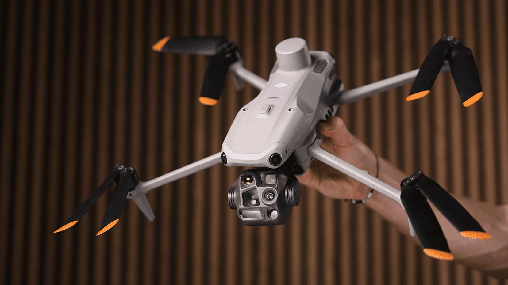 En person håller en avancerad DJI Matrice 4T-drönare med flera kameror och orange spetsar på propellrarna, mot en bakgrund av träpaneler.