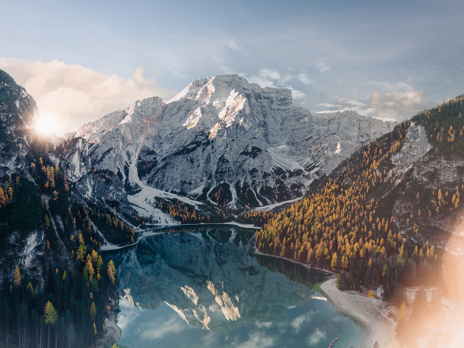 En soluppgång över ett bergslandskap med snötäckta toppar och en klar sjö omgiven av skog.