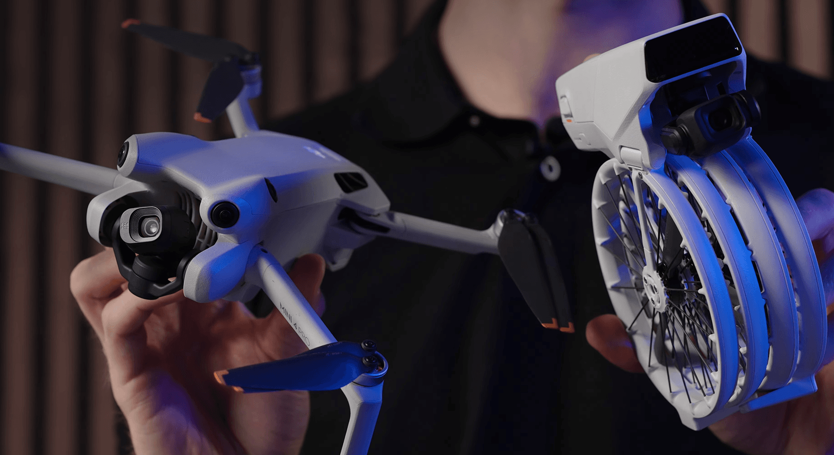 En person håller två avancerade drönare, en med utfällda propellrar och en kompakt hopfällbar modell, i en teknologisk miljö.
