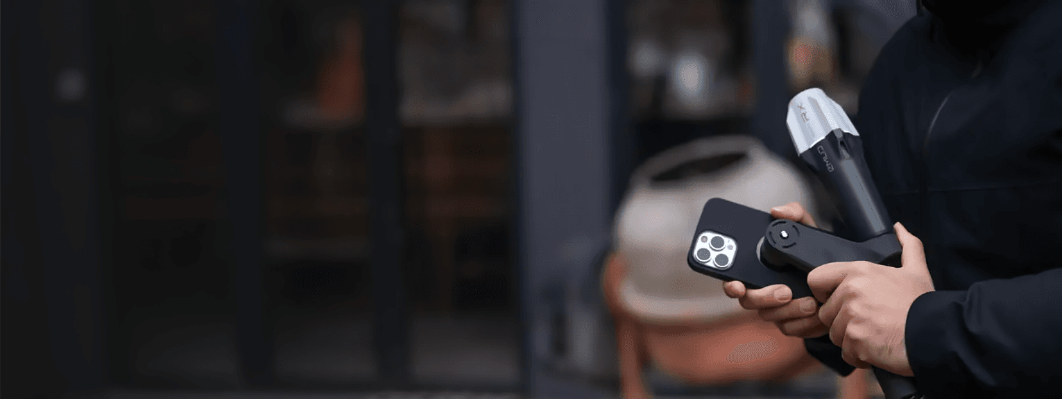 En person håller en smartphone och en Emlid Scanning Handle med Reach RX framför en suddig bakgrund av en byggnad.