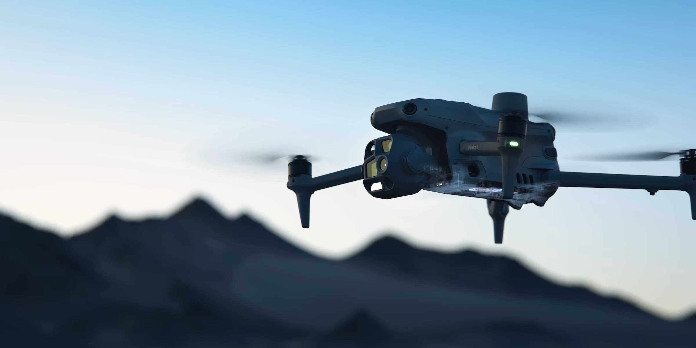 En DJI Matrice 4-drönare svävar i luften mot en bakgrund av bergstoppar i skymningen.