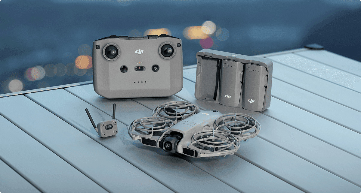 En DJI Neo 2-drönare med tillhörande fjärrkontroll och batterier är placerade på ett bord med en suddig stadsvy i bakgrunden.