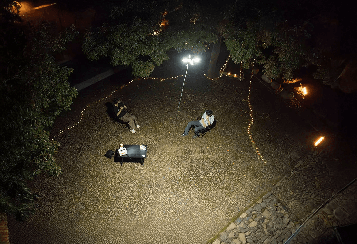 Två personer sitter på stolar i en upplyst av CZ10 Tethered Hover Light, omgivna av ljusslingor och träd, vilket skapar en intim atmosfär.