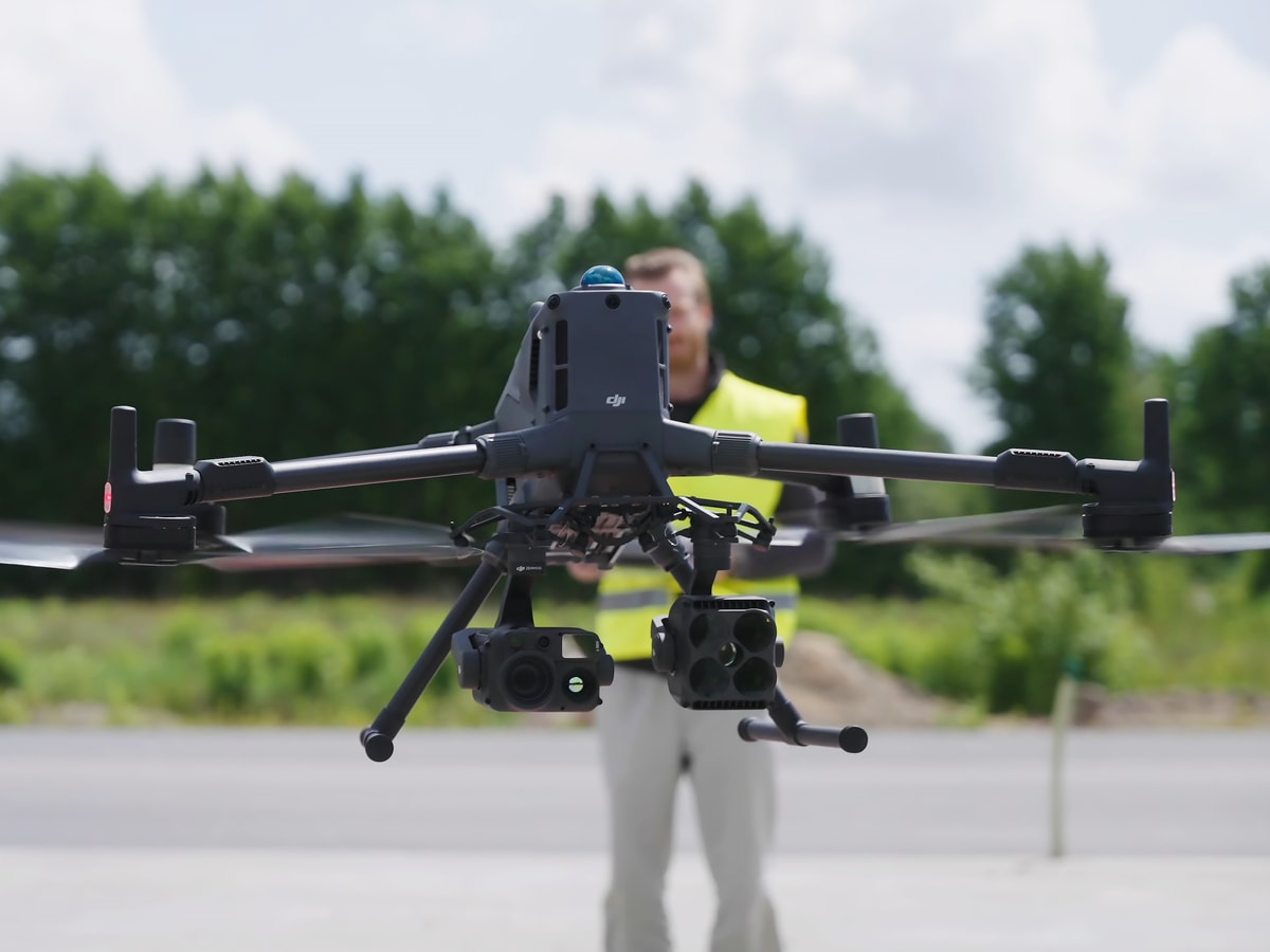 Drönare för inspektion: En DJI Matrice 400-drönare med en monterad DJI Zenmuse H30T-kamera svävar framför en person i en gul varselväst på en öppen plats med träd i bakgrunden.