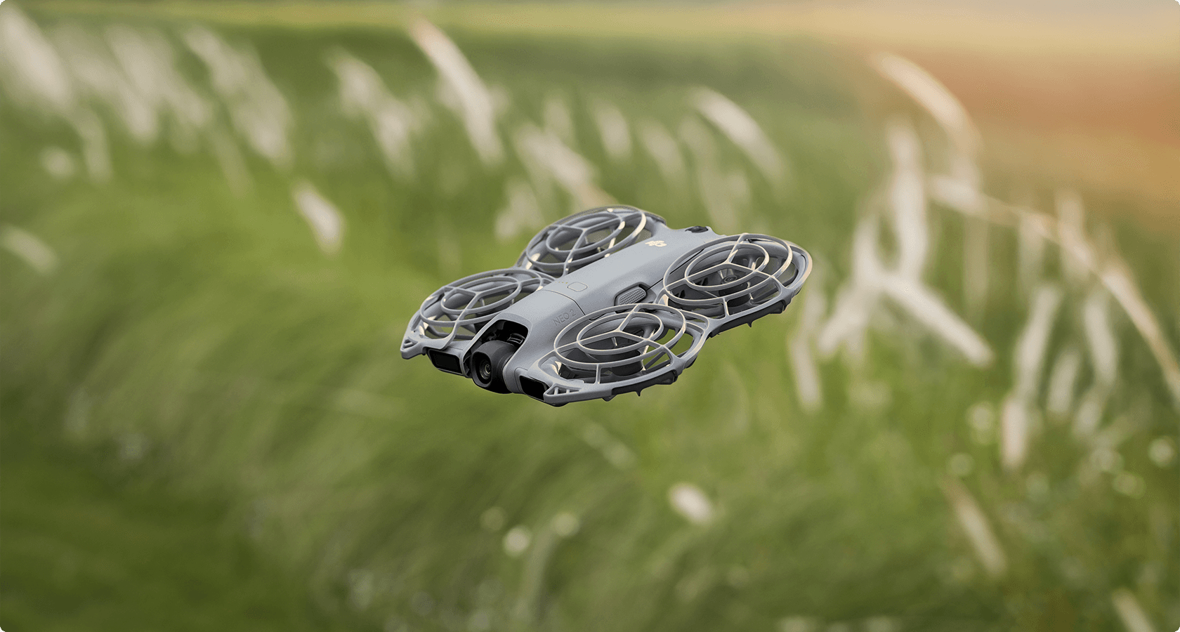En DJI Neo 2-drönare svävar över ett fält med högt gräs, fångad i en solig och naturlig miljö.