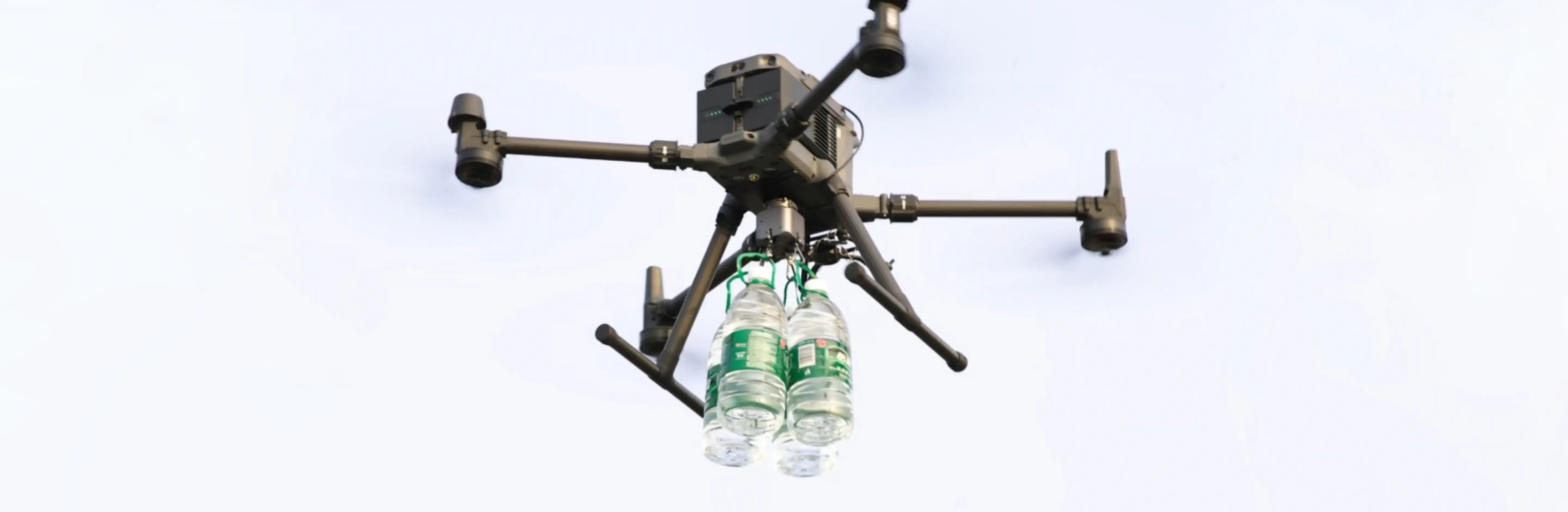 En drönare flyger genom luften och transporterar två vattenflaskor hängande under sig.